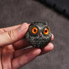 Une main tient une statue de hibou en résine artisanale aux yeux orange vif et aux plumes texturées.