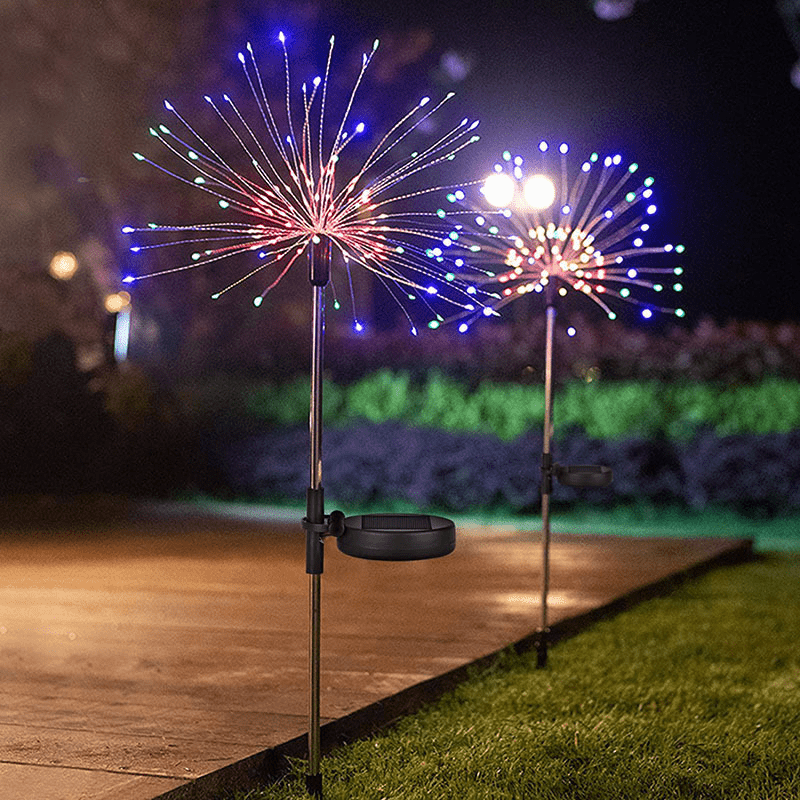 A tall metal stand holds a solar firework light display with bright blue and red sparks illuminating a dark outdoor garden.