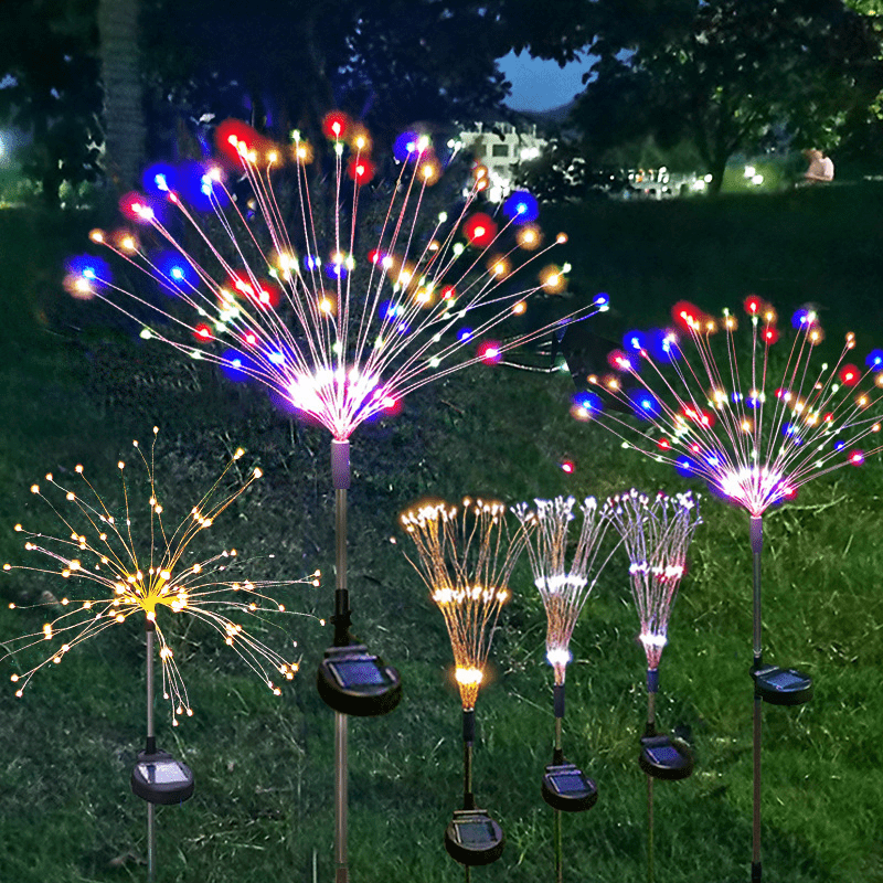 A display of bright, colorful solar fireworks lights shooting sparks into the night sky in a garden setting.