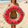 A woman holds a bright red inflatable pool float ring with black fruit designs on a sandy beach.