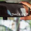 A hand is holding a rearview mirror sticker with the text Trust in God and a heart symbol.