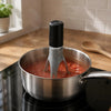A silver pot on a stovetop with an automatic pan stirrer inserted into the liquid.
