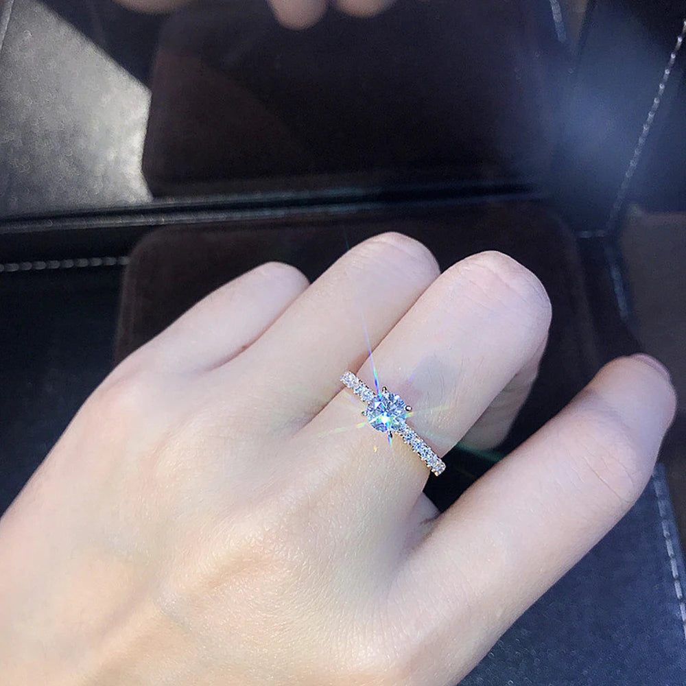 En hånd med en klassisk rund cubic zirconia-bryllupsring på ringfingeren, vist mot en mørk bakgrunn.