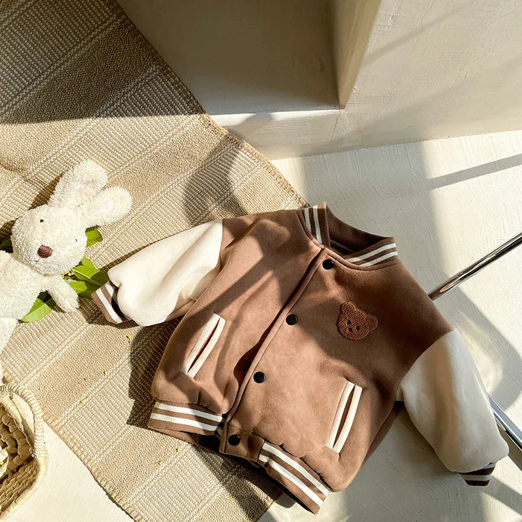 A brown corduroy jacket with white stripes and a brown patch is displayed on a woven mat next to a white stuffed animal.