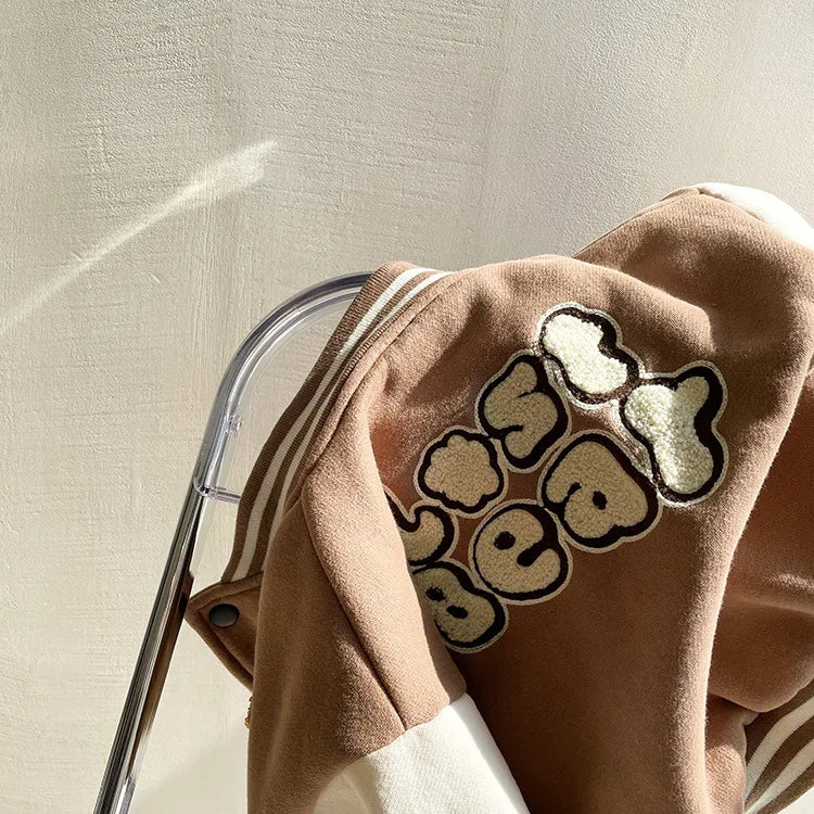 A close-up of a tan corduroy jacket with a beige bear graphic on the sleeve, shown on a hanger.