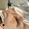 A close-up of a tan corduroy jacket featuring a round, textured, light brown bear appliqué on the chest.