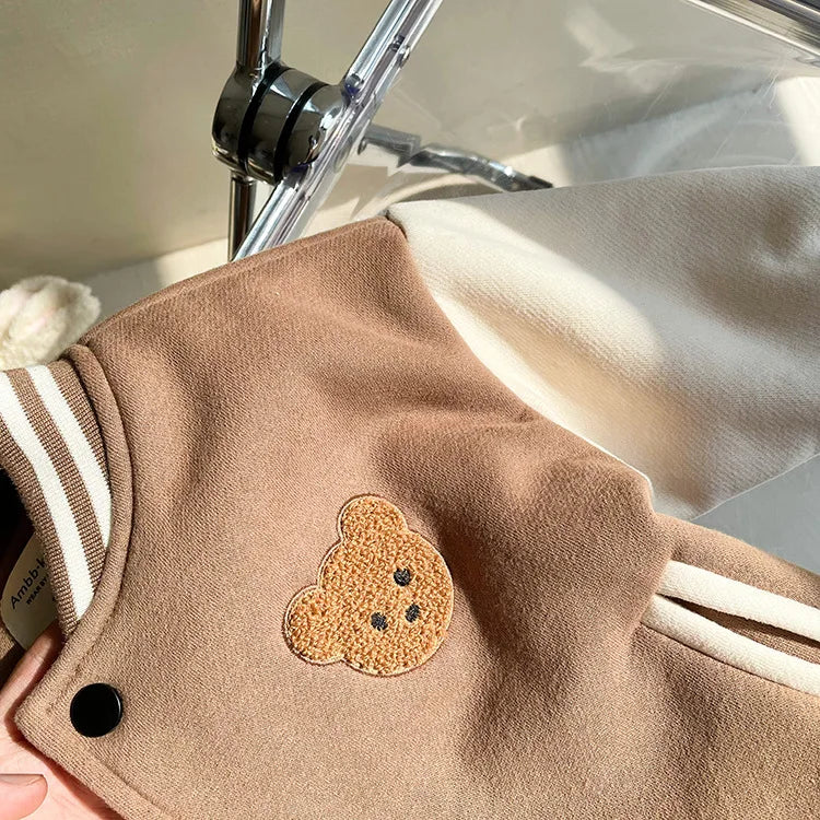 A close-up of a tan corduroy jacket featuring a round, textured, light brown bear appliqué on the chest.