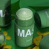 A green cleansing stick mask product in a glass jar is shown on a green surface with yellow leaves.