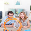 A family is shown playing with an orange and blue DIY stunt car racing track set and launchers on a wooden table.