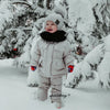 A baby wearing a beige winter down jacket and pants set with a faux fur hood standing in the snow.