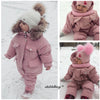 A baby wearing a pink winter down jacket and pants set with a faux fur hood standing in the snow.