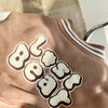 A close-up of a tan fleece jacket with embroidered beige lettering on the chest.