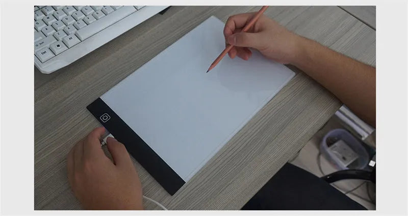 يد ترسم على لوح رسم LED أبيض مقاس A4 بقلم رصاص على مكتب خشبي