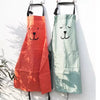 An orange cartoon bear cotton apron hanging on a white wall next to a green one.