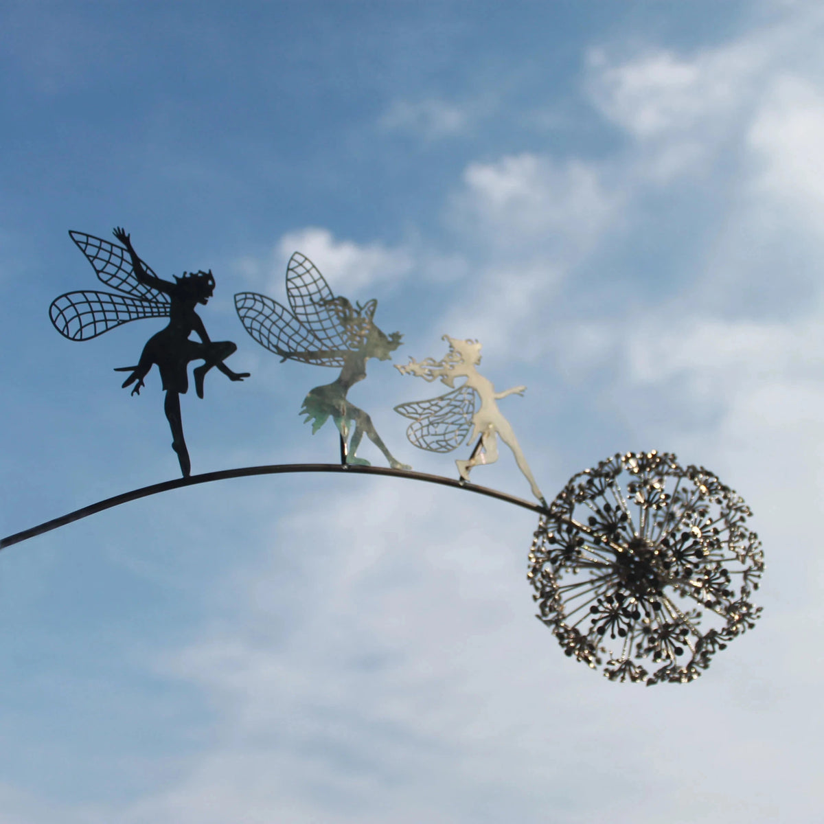 En metallskulptur av en fe med vinger som står på en tråd, ved en løvetannfrøstand mot en blå himmel.