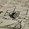 A small, dark green mini 3x3 speed cube resting on a sheet of scientific or mathematical paper.