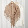 A beige knitted cotton tassel wrap shawl displayed on a wooden hanger against a white background.