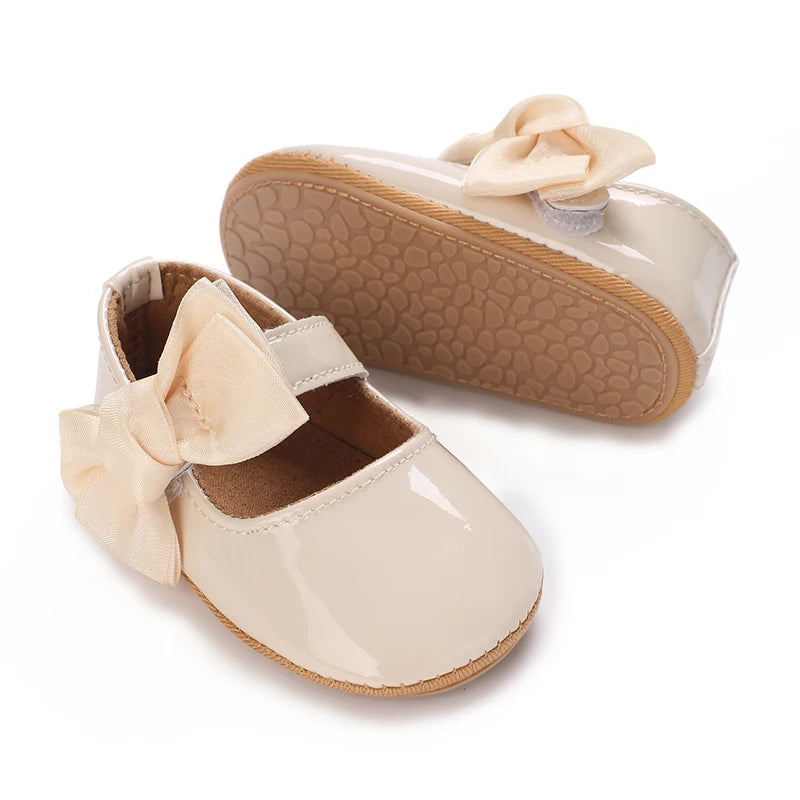 A pair of soft beige baby first walker shoes with a light pink bow and a textured sole.