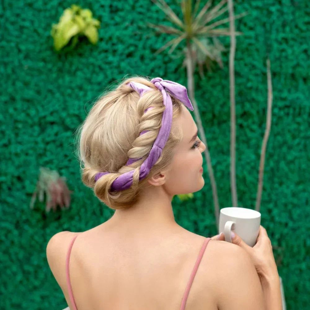 A woman with blonde hair wearing a purple heatless curling rod headband, posing against a green textured wall.