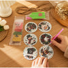 A hand is assembling a festive diamond painting wooden ornament kit with colorful beads and a pink tool on a wooden surface.