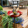 A handcrafted metal bird bath in a terracotta-colored bowl is placed in a wooden garden planter surrounded by green foliage a
