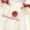 A cream-colored kids summer pinafore dress featuring a strawberry embroidery and a red plaid bow detail.