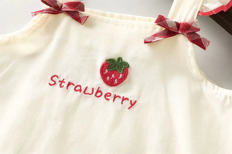 A cream-colored kids summer pinafore dress featuring a strawberry embroidery and a red plaid bow detail.