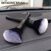 A black detailing brush with a silver ferrule and a fluffy, gray-white synthetic hair is shown on a dark surface.