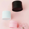 Two refillable travel face cream jars in black and white are displayed on a pink background.