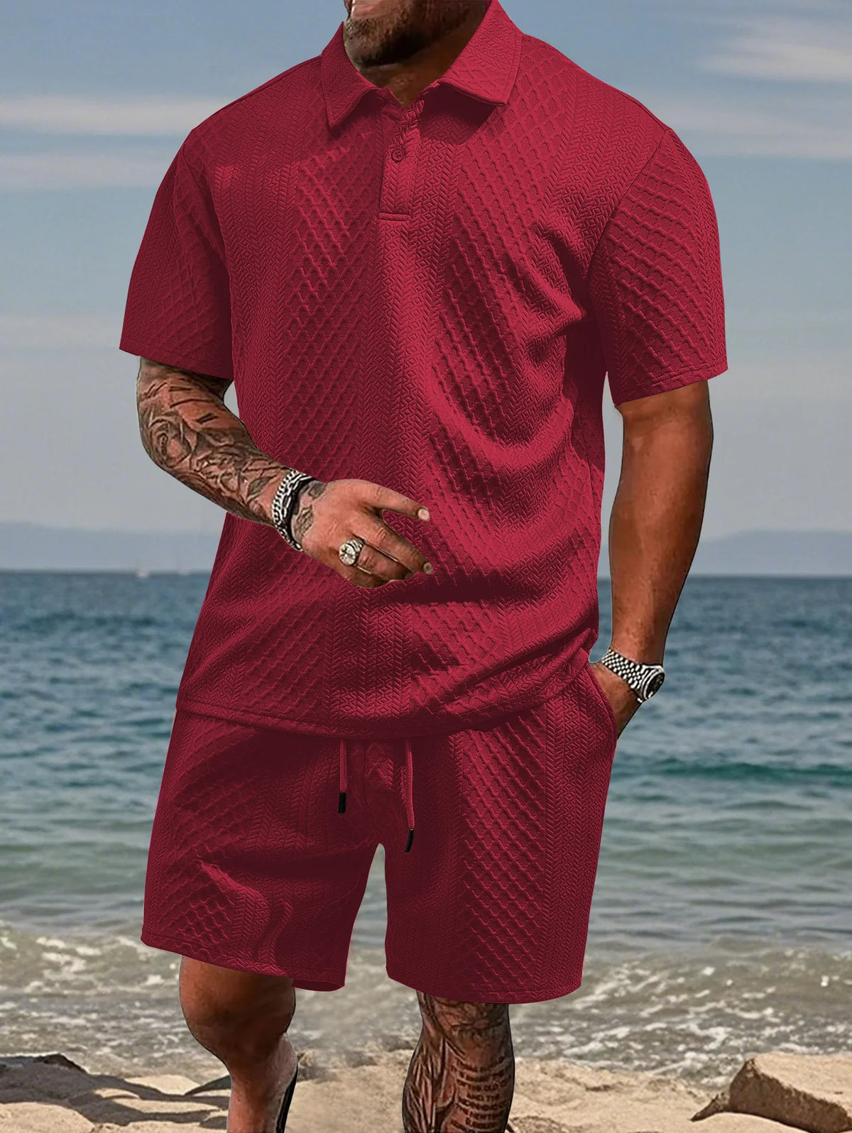 A man wearing a maroon jacquard short-sleeve shirt and matching drawstring shorts set standing near the ocean.