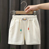 Cream kids beach shorts with elastic waist featuring a small embroidered logo and colorful tassels