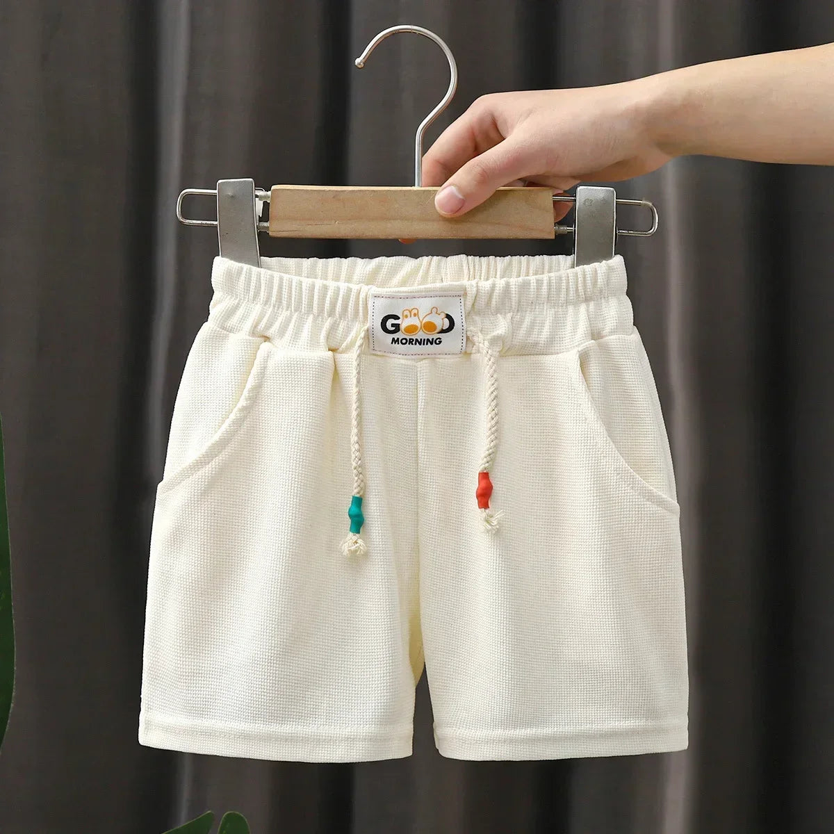 Cream kids beach shorts with elastic waist featuring a small embroidered logo and colorful tassels