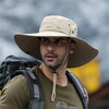 A man wearing a beige wide brim mesh bucket hat and olive green shirt, carrying a backpack, outdoors.