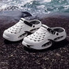 White men's EVA slip-on sandals with cushioned platform soles resting on dark rocks near the sea.