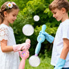A girl and a boy are playing with a pink smoke bubble wand and clear bubbles outdoors.