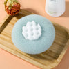 A light blue exfoliating bath sponge with a textured surface is placed on a wooden tray next to a white container.