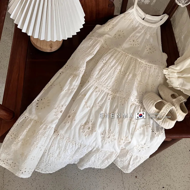 A white cotton suspenders dress for a toddler girl is displayed on a wooden surface.