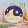 Bordado nocturno de luna morada y amarilla en lienzo redondo rodeado de herramientas metálicas doradas.