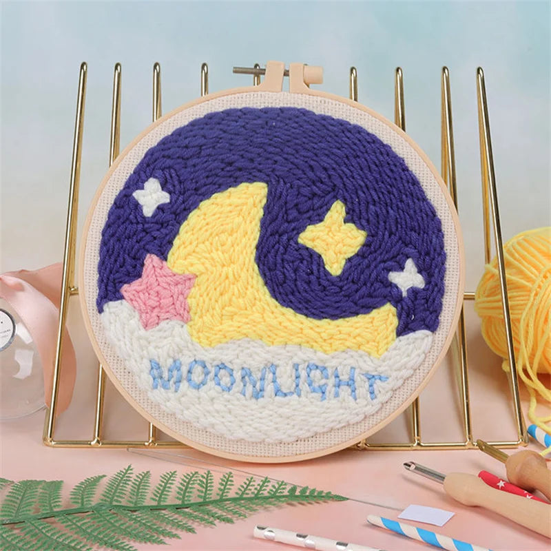 Bordado nocturno de luna morada y amarilla en lienzo redondo rodeado de herramientas metálicas doradas.