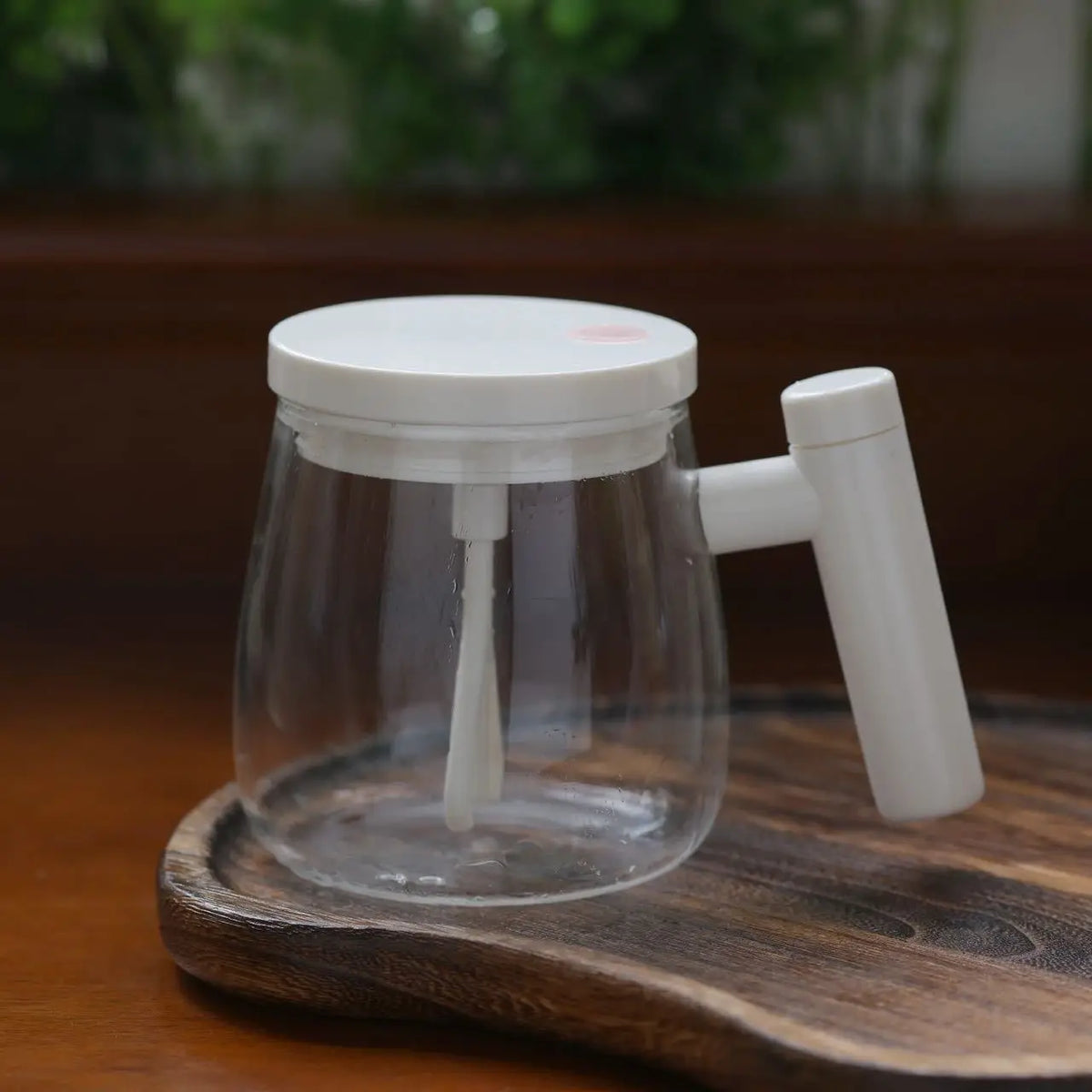 A clear glass mug with a white lid and a white self-stirring mechanism and wooden handle resting on a dark cutting board.