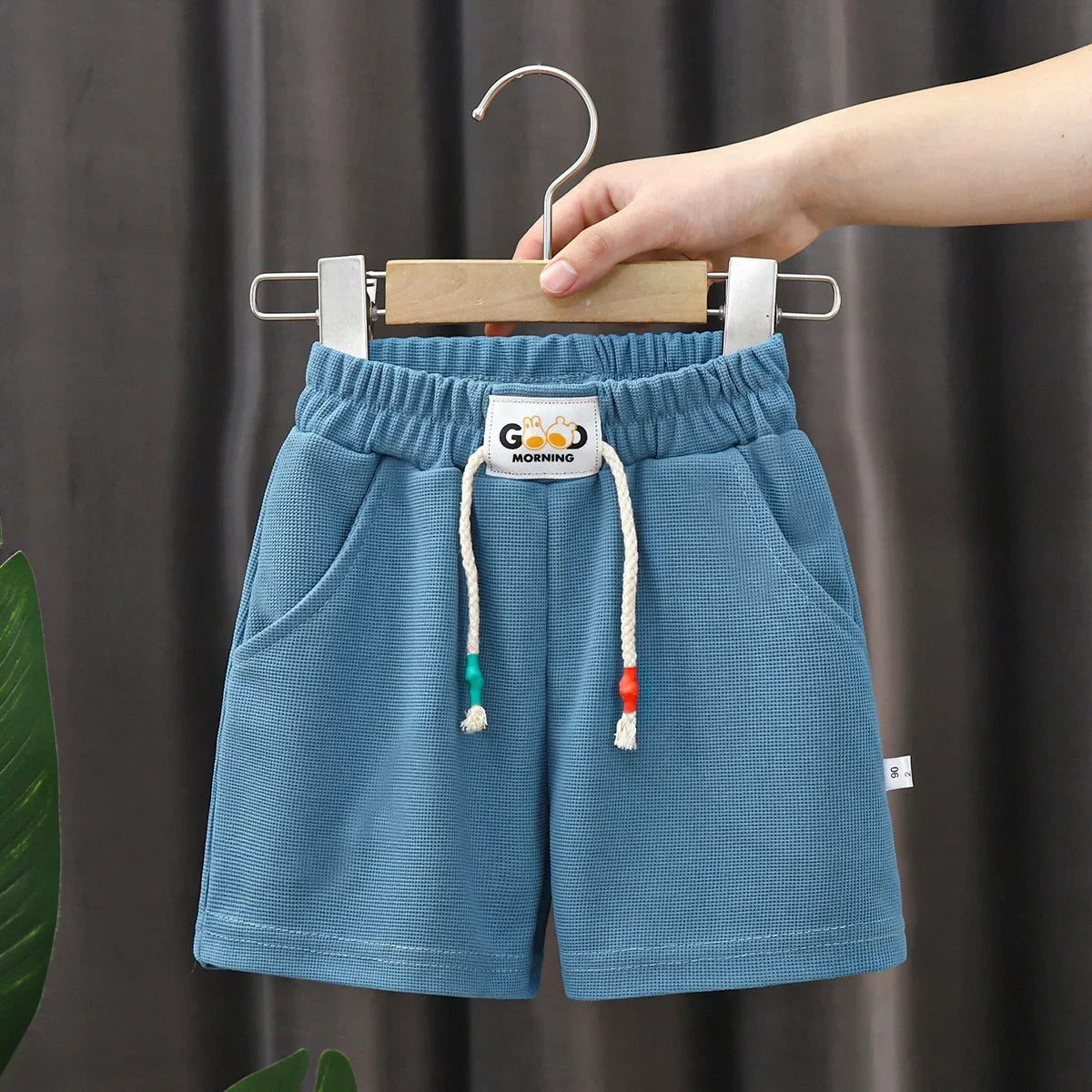 Light blue kids beach shorts with an elastic waist and cord drawstring hanging on a wooden hanger