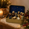 A classic nativity scene in a gold-colored metal tin box featuring figures of Mary, Joseph, and the baby Jesus under a starry