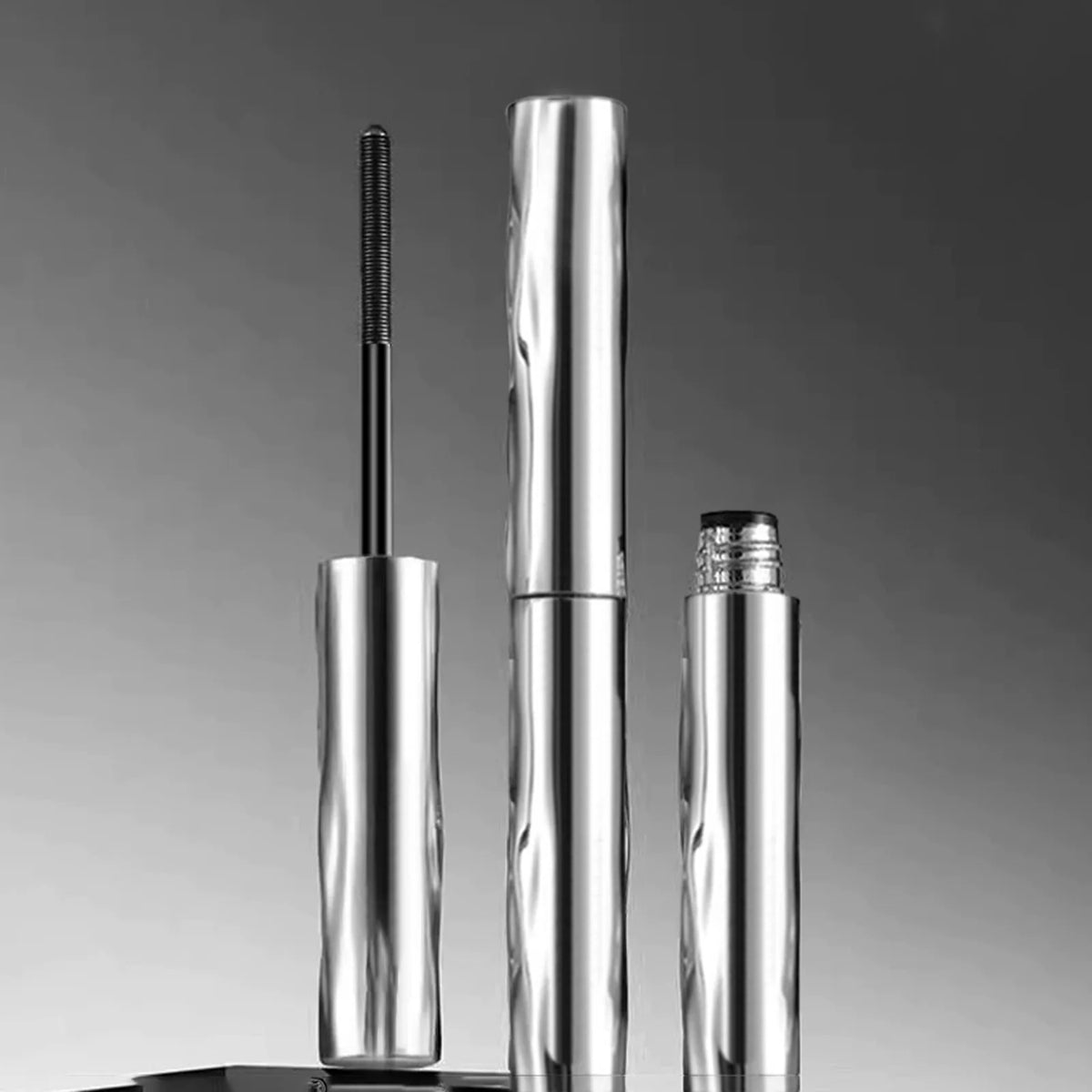 A silver waterproof volumizing mascara and defining brush standing vertically against a gray background.