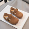 A pair of tan suede non-slip kids' moccasin shoes with a hook-and-loop closure resting on a white card.