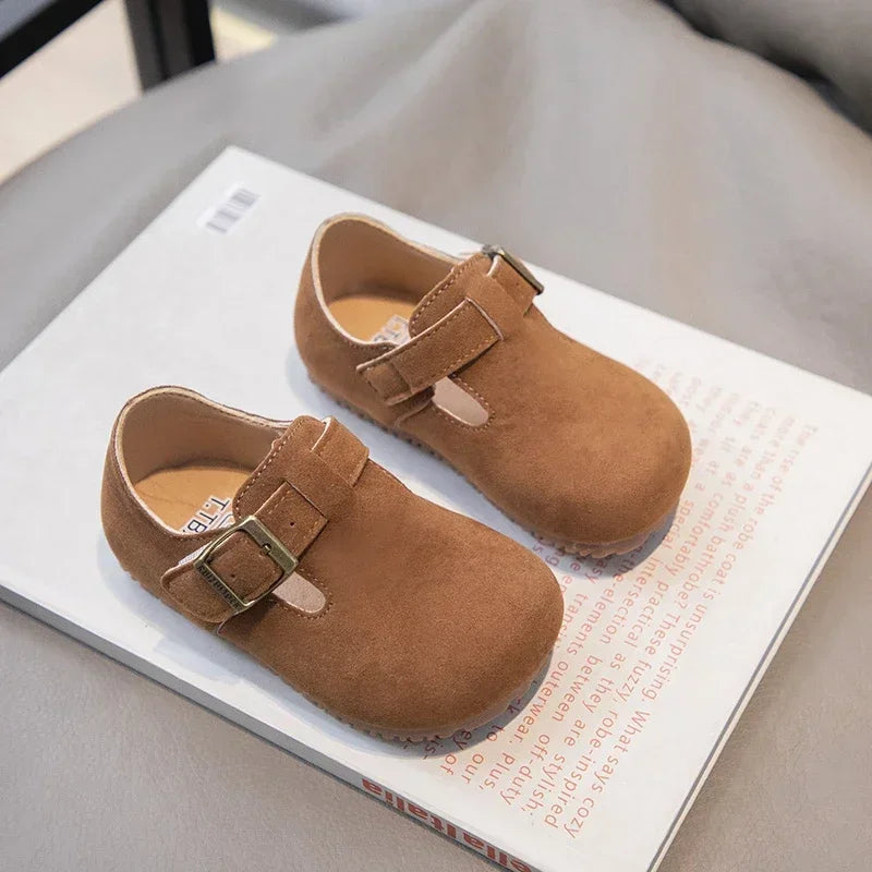 A pair of tan suede non-slip kids' moccasin shoes with a hook-and-loop closure resting on a white card.