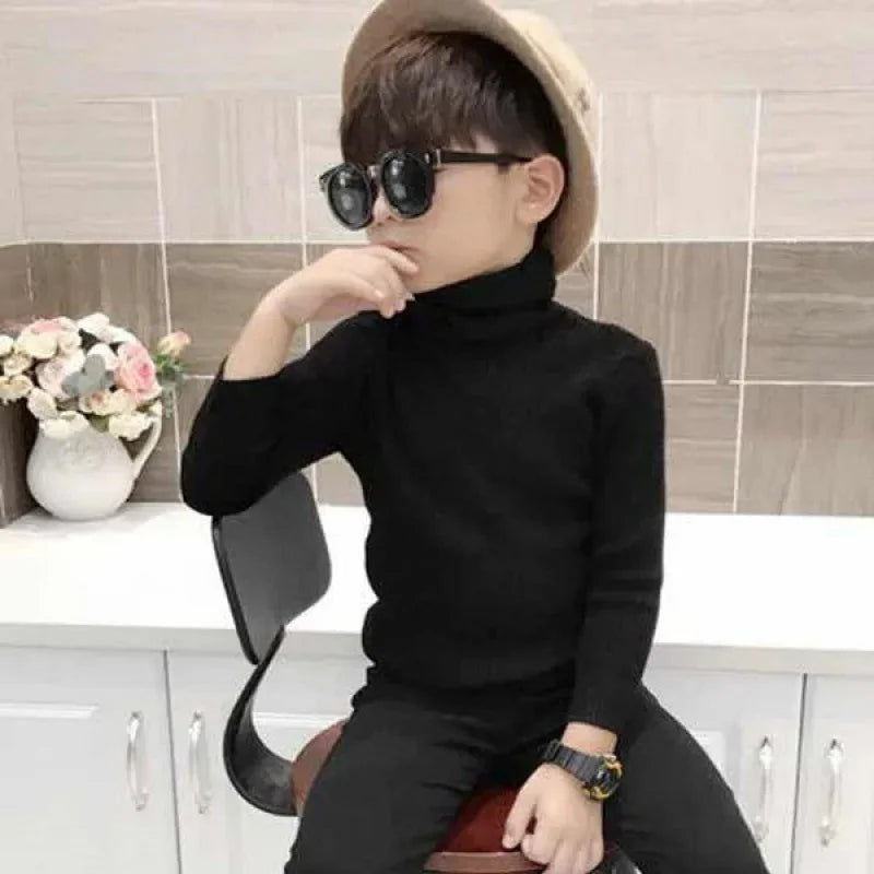 A young boy wearing a black turtleneck sweater and sunglasses is seated in front of a white cabinet.