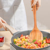 A hand is using a natural teak wood cooking spatula to stir shrimp and vegetables in a pan.