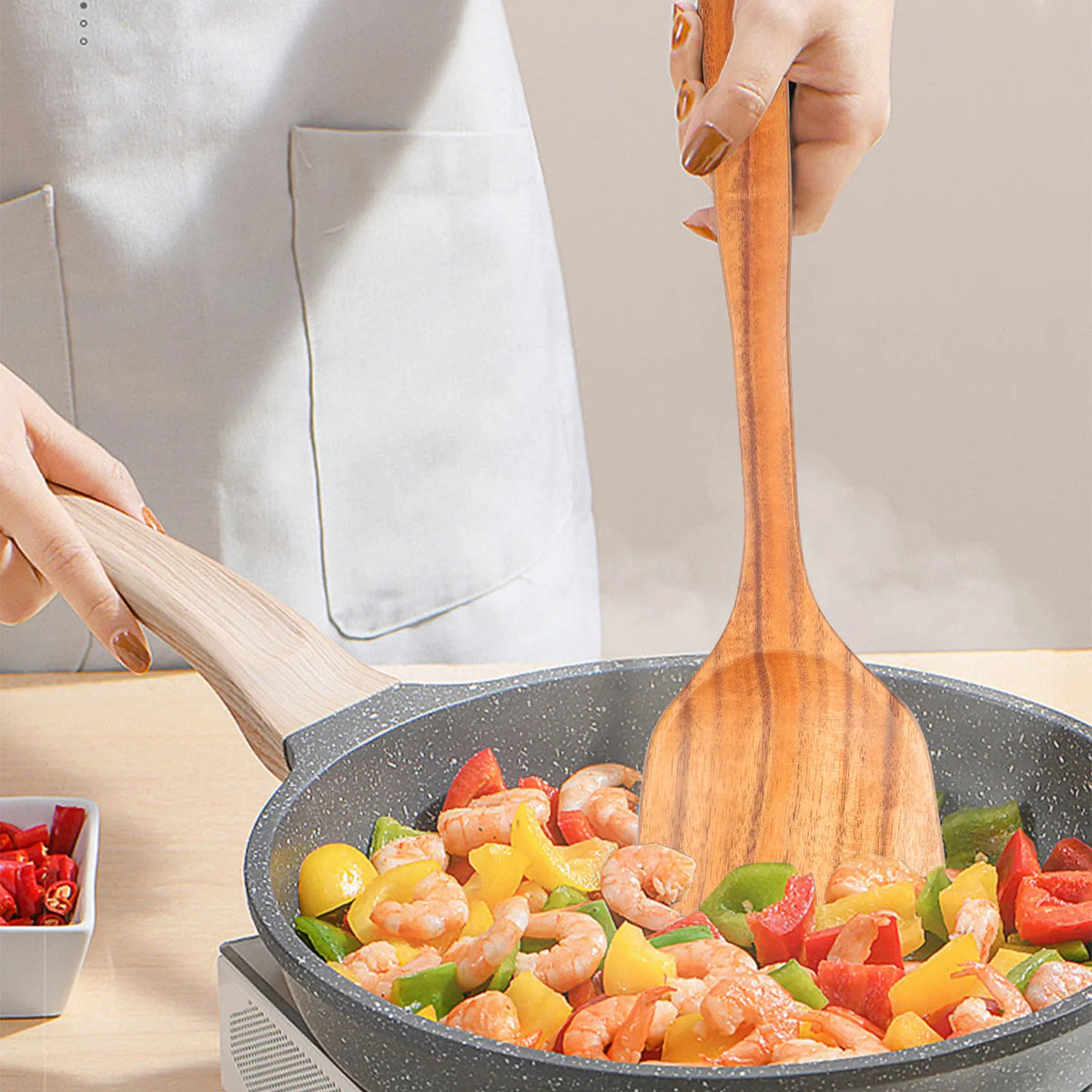 A hand is using a natural teak wood cooking spatula to stir shrimp and vegetables in a pan.
