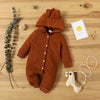 A brown knitted hooded baby romper lying on a wooden surface with a sprig of greenery and a yellow smiley card.