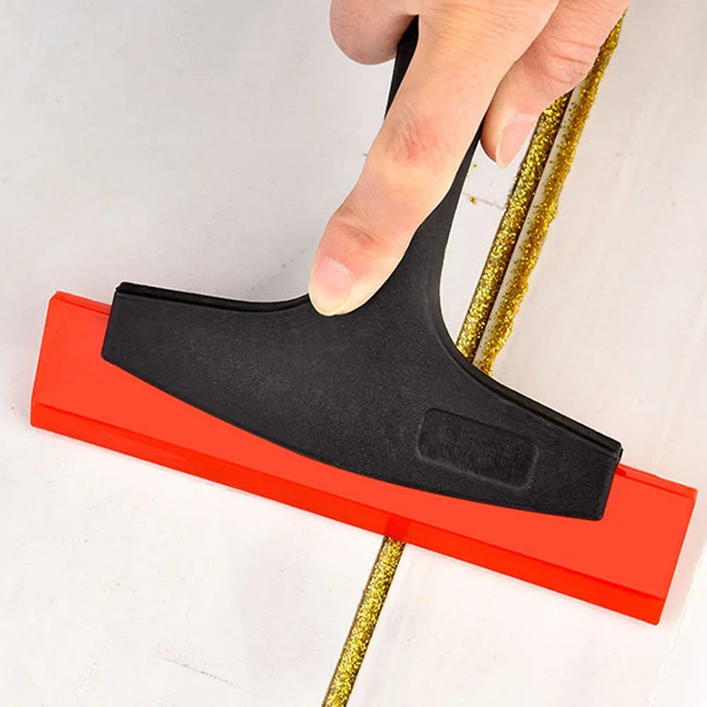 A hand is using a black tile gap filling and cleaning scraper set with a red plastic base and a gold wire tool.